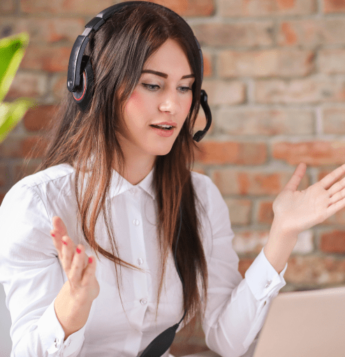 Woman with long brown hair wearing a white blouse and headset, gesturing with her hands while talking about CTA compliance now, against a brick wall background.