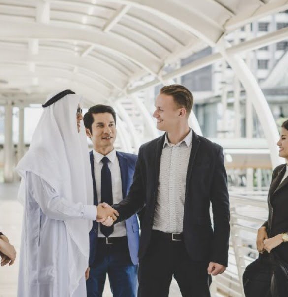 A group of business professionals, including one in traditional Middle Eastern attire, stand in an atrium while two men in suits shake hands, smiling and talking about the Corporate Transparency Act.