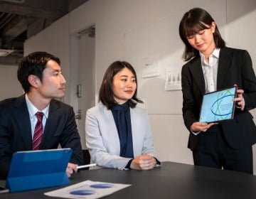 Three people in business attire are in a meeting room. One person is standing and showing a tablet to two seated individuals, decoding the details of the Corporate Transparency Act. Documents and another digital device are on the table, offering further guidance for businesses.