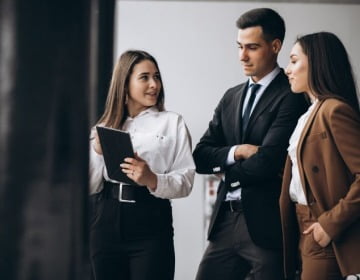 Three individuals in professional attire are having a discussion indoors. One holds a tablet and appears to be explaining something about beneficial ownership reporting to the other two. All appear engaged in navigating the complexities of this practical guide.