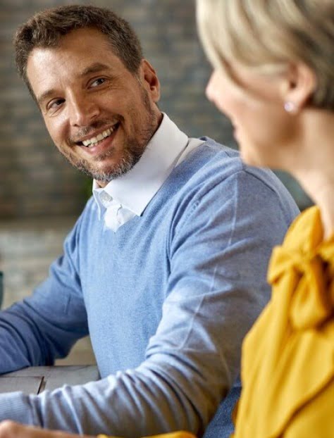 A man wearing a blue sweater and white shirt smiles at a woman in a yellow blouse while conversing, perfectly capturing the warm interactions our "About Us" page embodies.