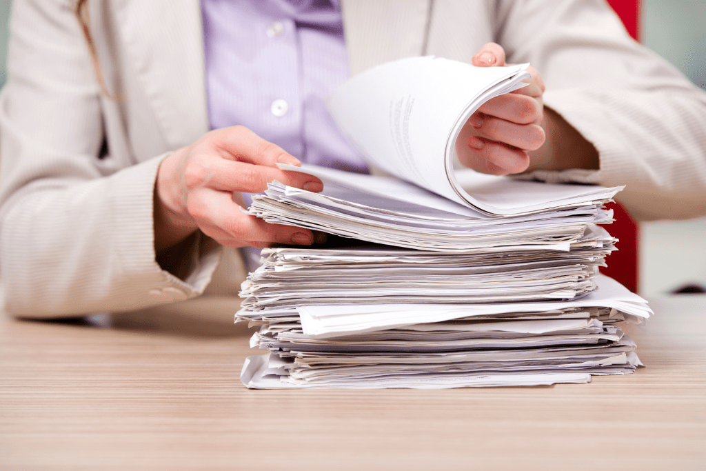 A person in a beige suit reviews a large stack of papers on a desk, meticulously working to create a compliance strategy.