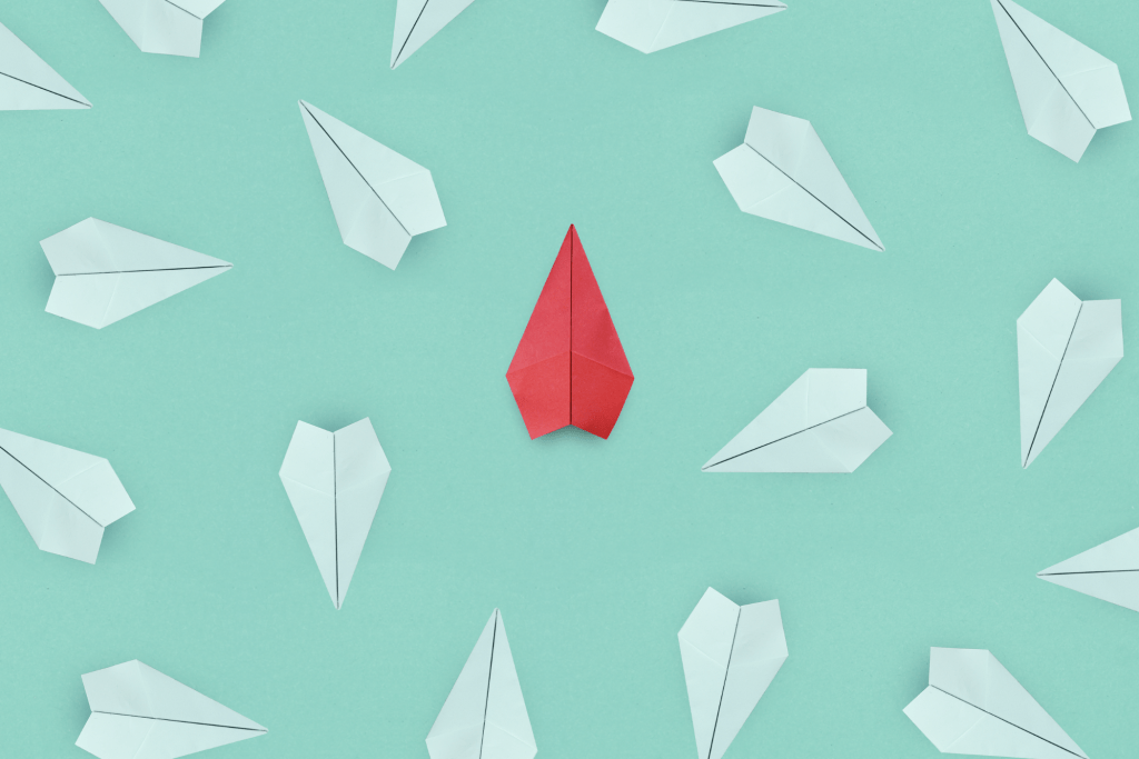 Origami paper airplanes in white scattered around a single red paper airplane on a light teal background illustrate the business advantage for SMBs striving for compliance.