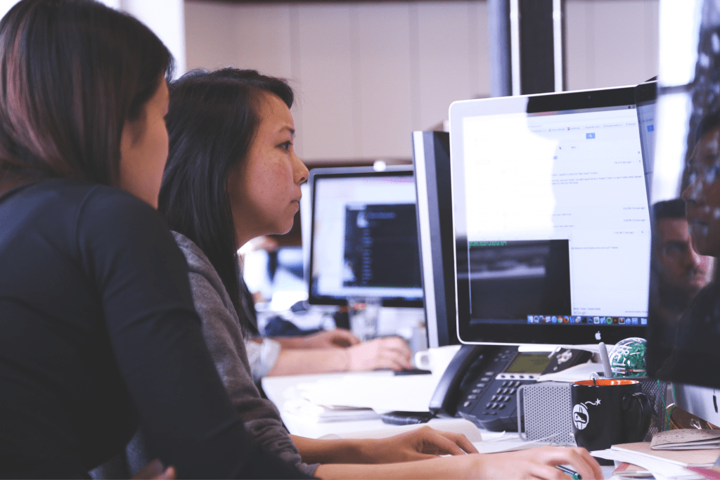 Two women are working together at a desk with computers, both intently focused on their screens. An office environment buzzes in the background as they collaborate on developing a compliance program.