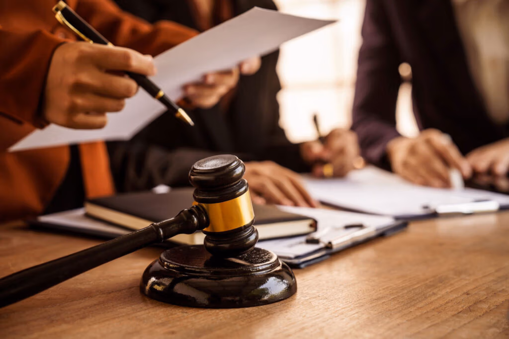 A wooden judge's gavel rests on a desk while three people review and sign documents in the background, preparing for new CTA filing requirements set to take effect in May 2025.